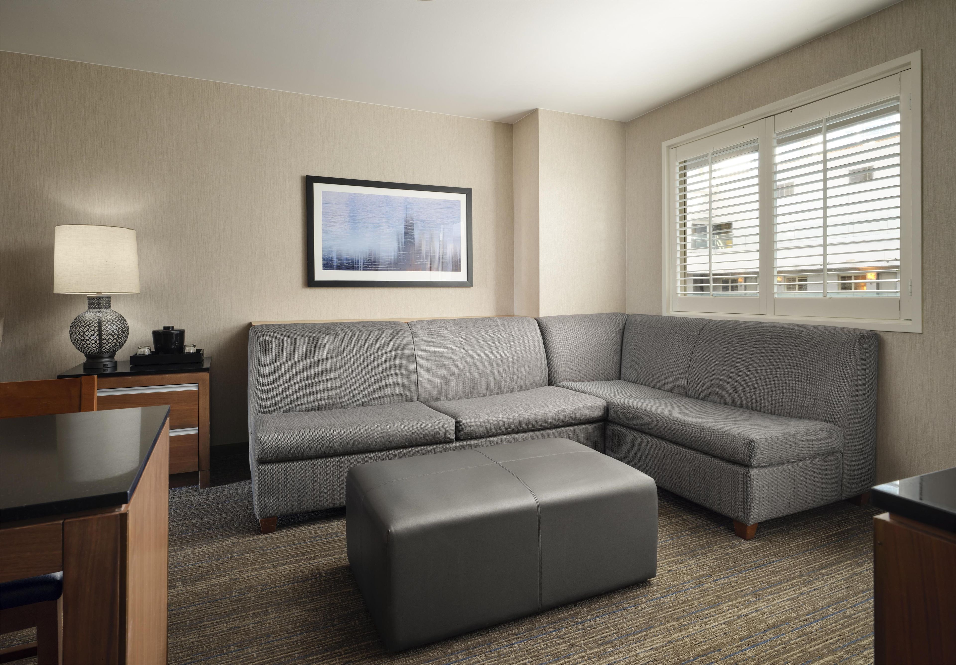 A modern hotel room with a gray sectional sofa, a small ottoman, a side table with a lamp, and a framed cityscape picture on the wall.
