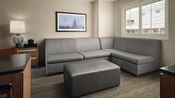 A modern hotel room with a gray sectional sofa, a small ottoman, a side table with a lamp, and a framed cityscape picture on the wall.