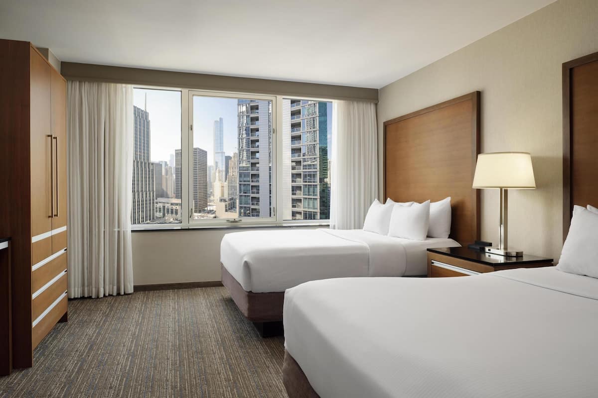 A hotel room with two beds, a large window with a city view, a wooden wardrobe, and a bedside table with a lamp.