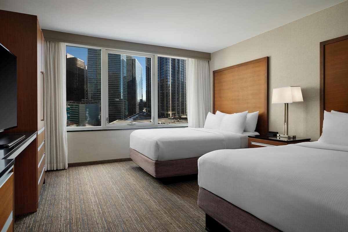 A hotel room with two beds, a television, a lamp, and a view of tall buildings.