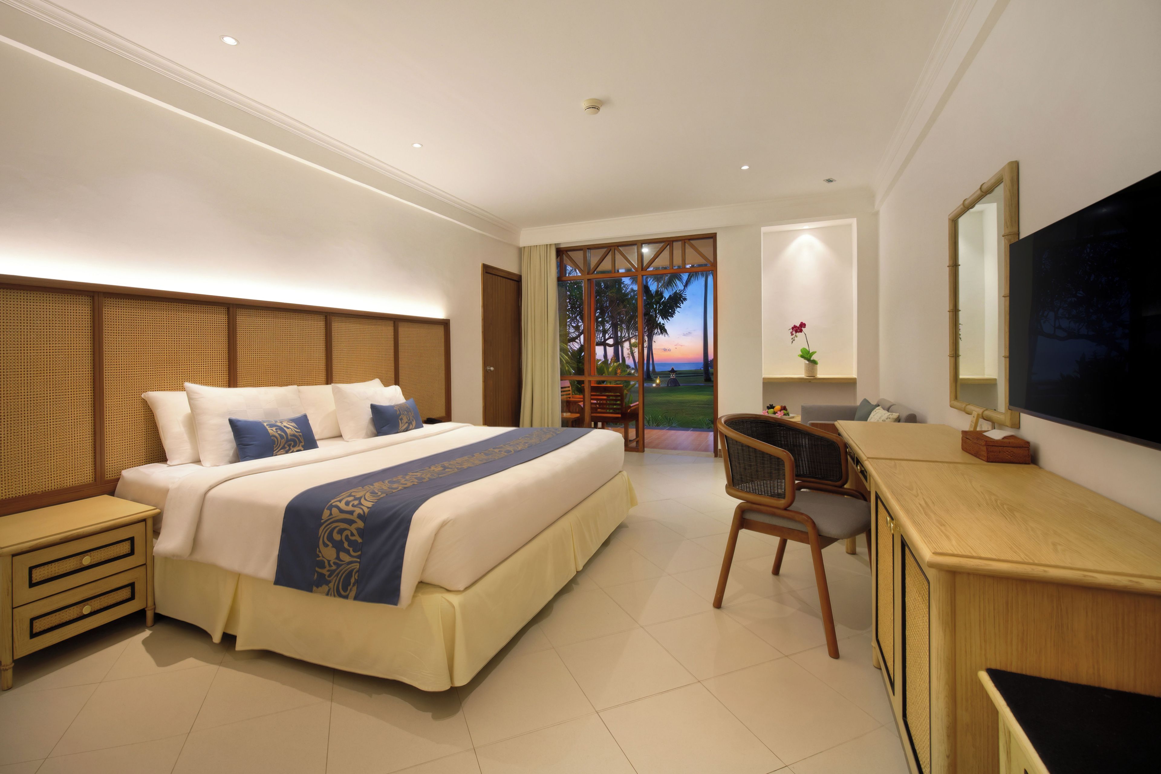 Ein Hotelzimmer mit einem großen Bett, einem Schreibtisch und Blick auf Palmen durch eine Tür.