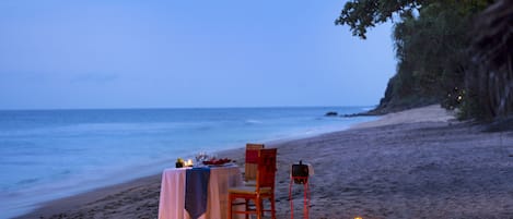Mesa junto a la playa dispuesta con velas, un mantel y una silla, con vista al océano.