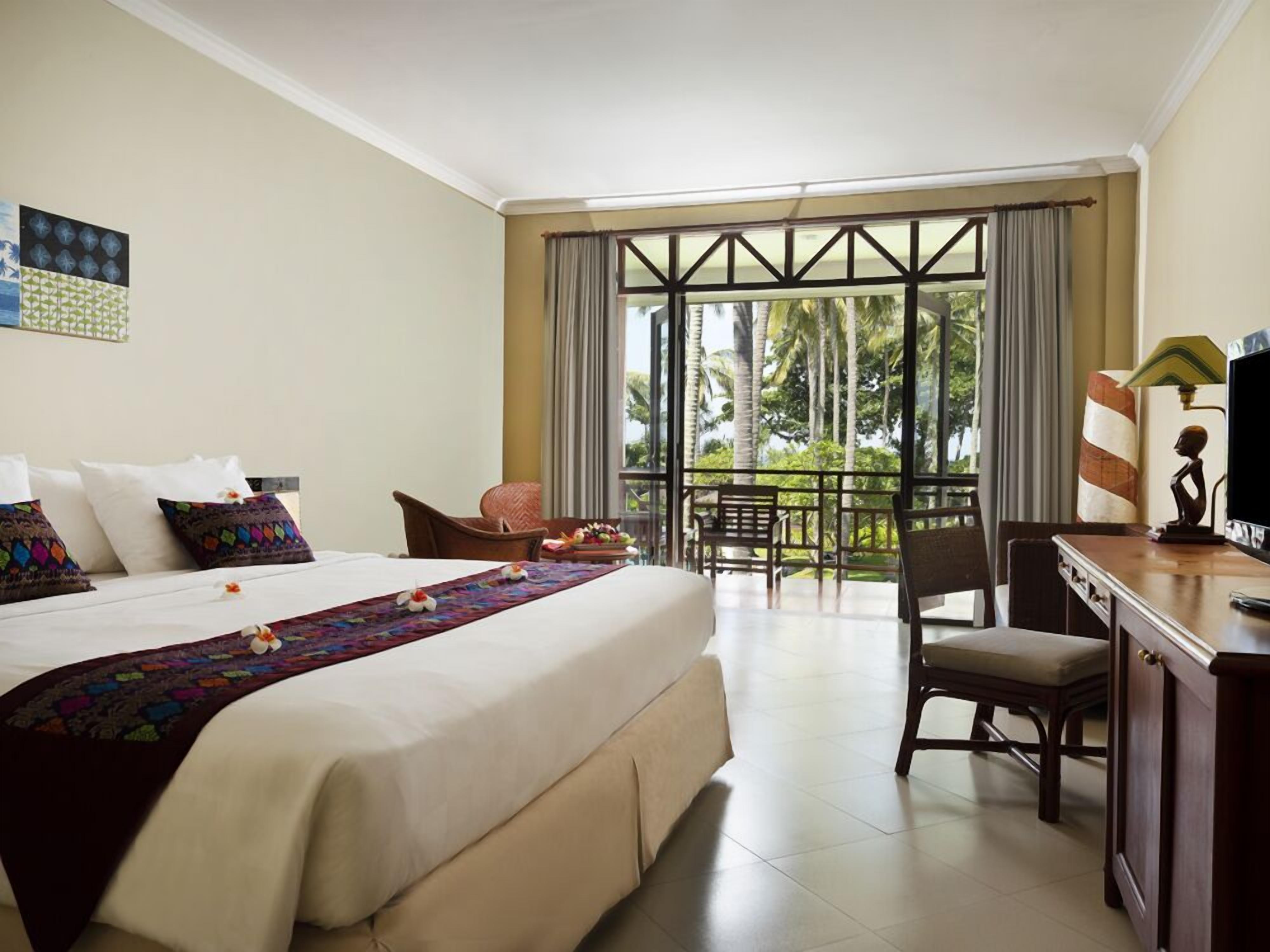 Een hotelkamer met een groot bed, een bureau, een stoel en een balkon met uitzicht op palmbomen.