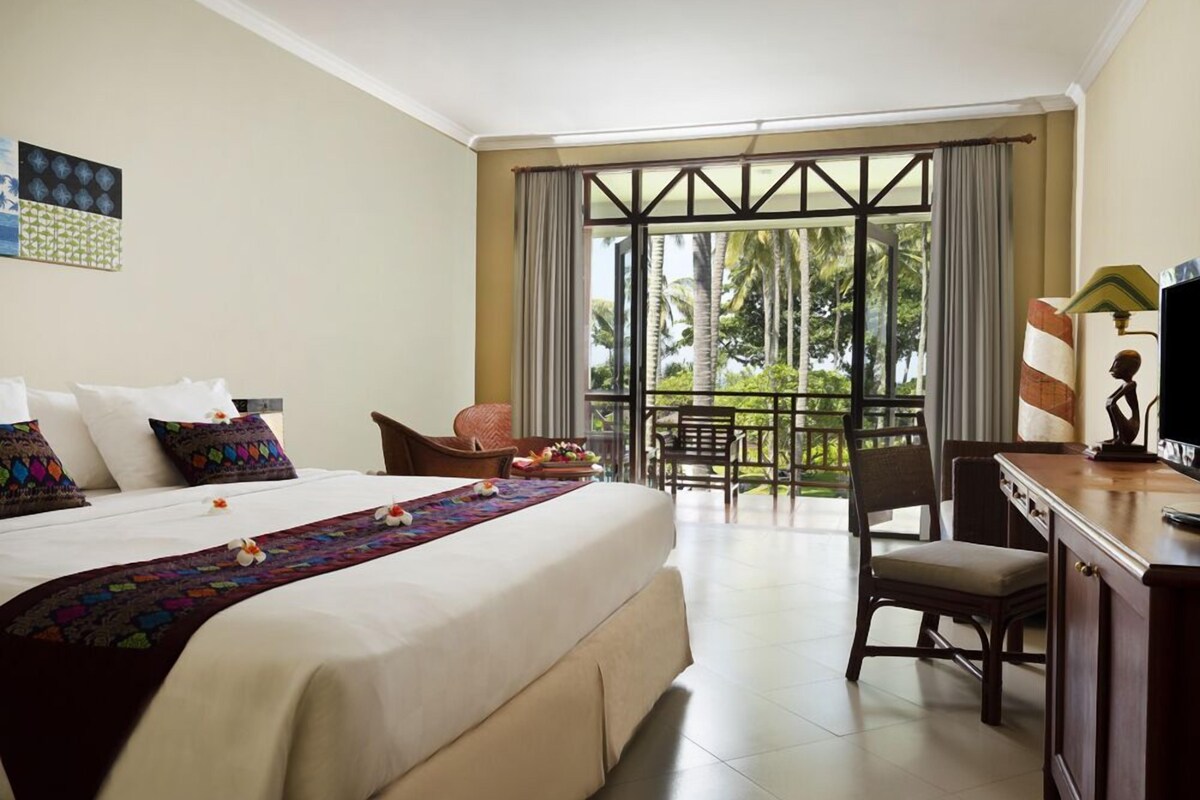 A hotel room with a large bed, a desk, a chair, and a balcony with a view of palm trees.