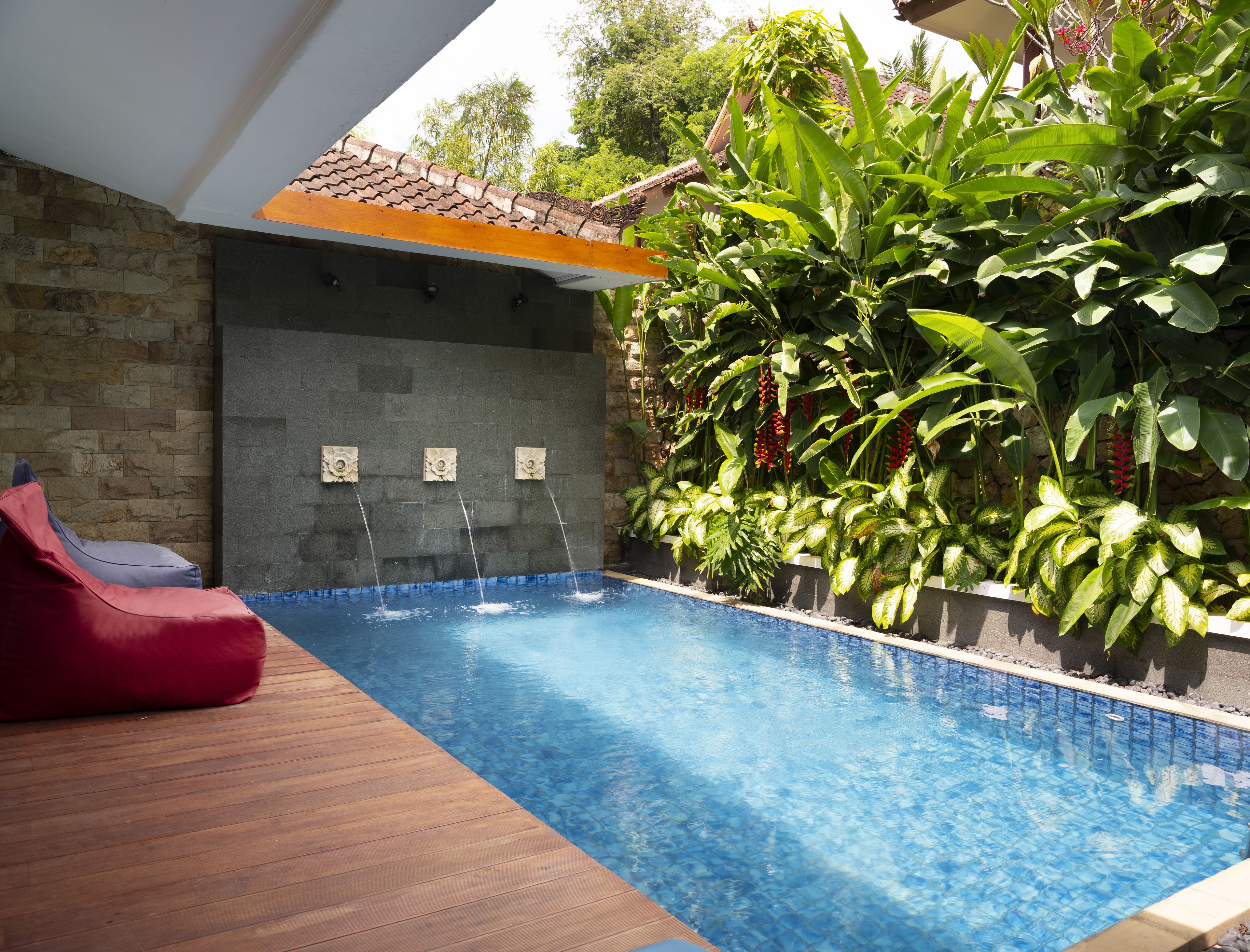 A swimming pool with a wooden deck, surrounded by lush greenery and a stone wall.