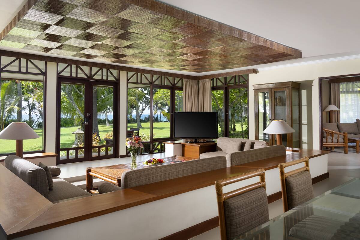 A spacious living room with a large TV, comfortable seating, and a view of a lush garden and ocean.
