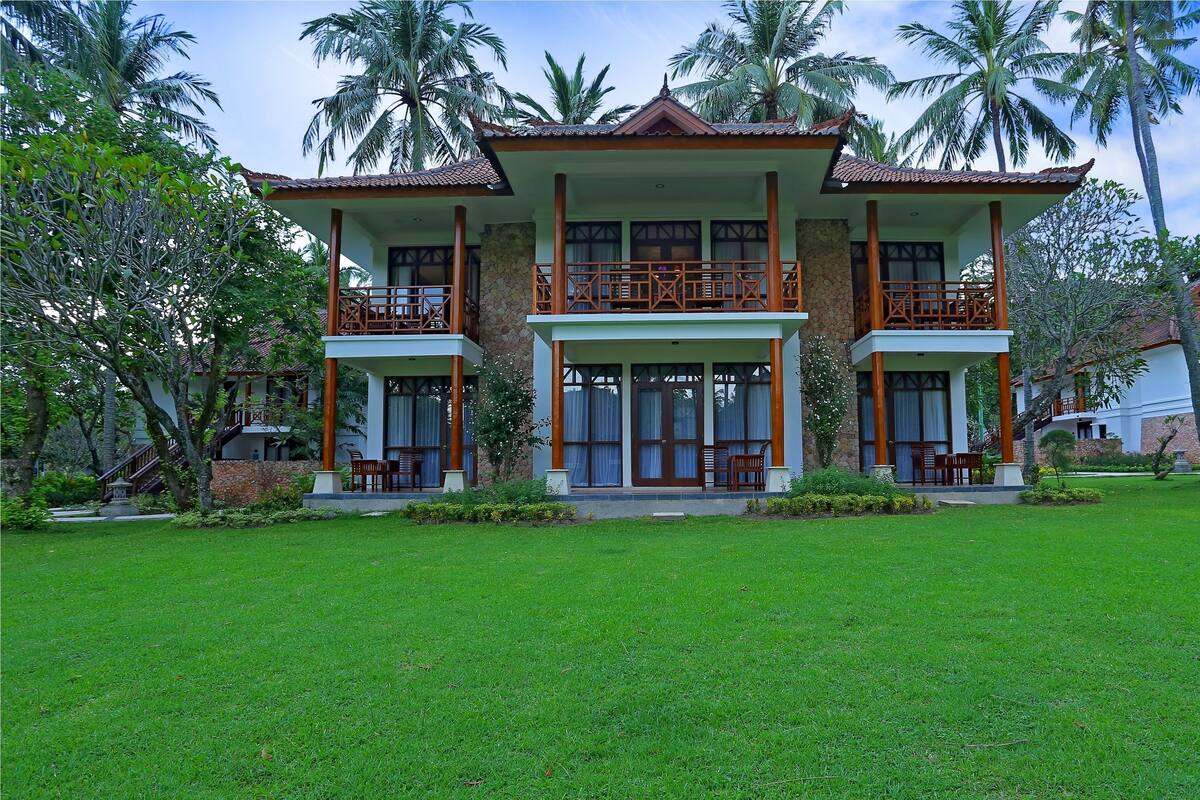 A two-story building with a balcony on each floor, surrounded by lush greenery and palm trees.