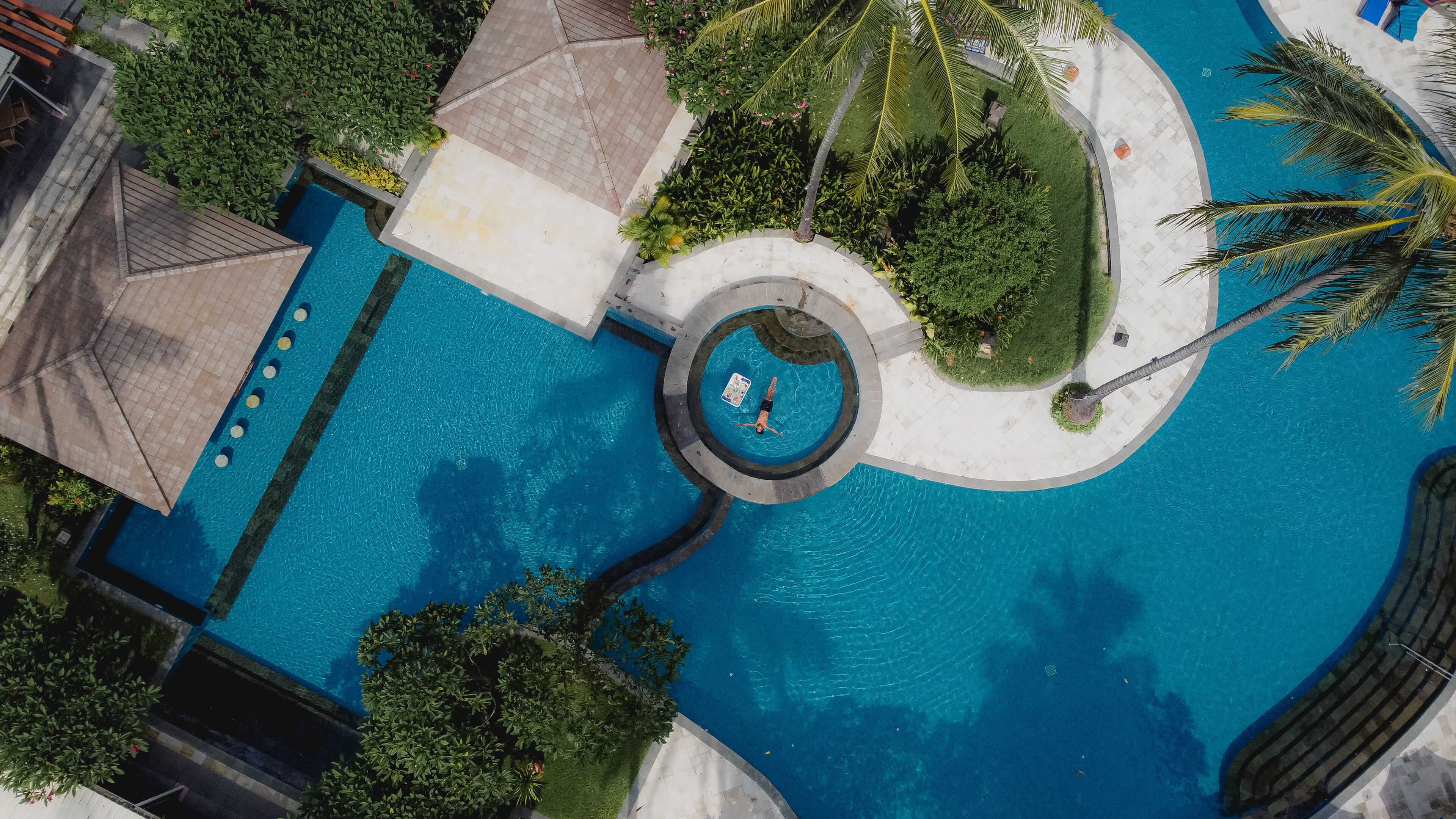 An aerial view of a swimming pool with a central circular feature, surrounded by palm trees and greenery.