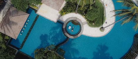 An aerial view of a swimming pool with a central circular feature, surrounded by palm trees and greenery.