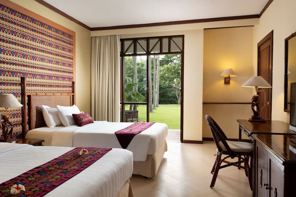 A hotel room with two beds, a desk, a chair, a large window with a view of greenery, and a patterned wall.
