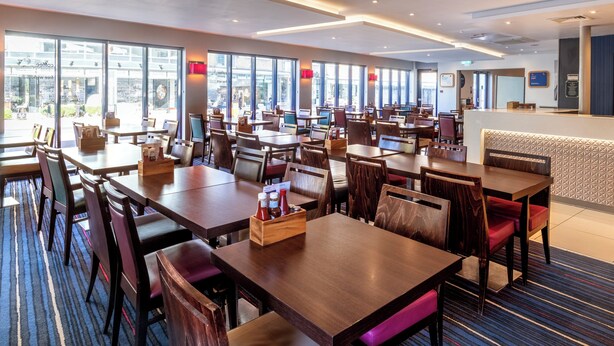 A modern hotel dining area with multiple tables and chairs, large windows, and a reception desk. - Holiday Inn Express London Wandsworth by IHG (London)
