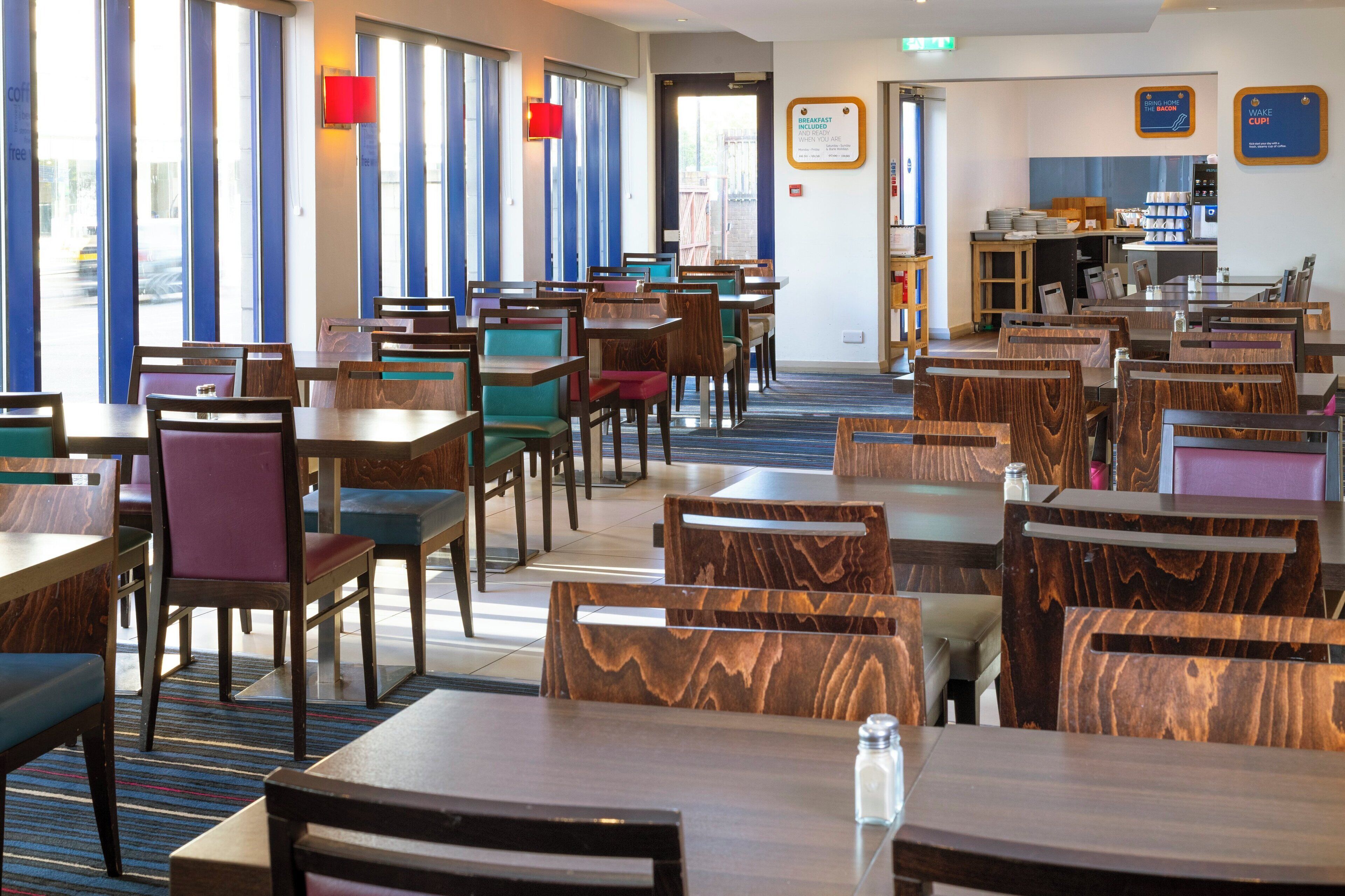 A hotel dining area with multiple tables and chairs, a breakfast counter, and a view of the street through large windows.