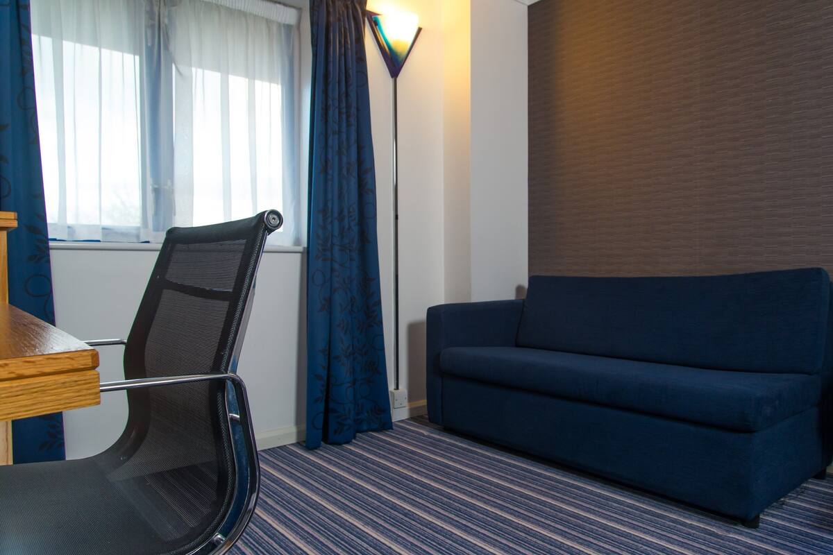 Une pièce avec un canapé bleu, un fauteuil, une lampe et une fenêtre avec des rideaux.