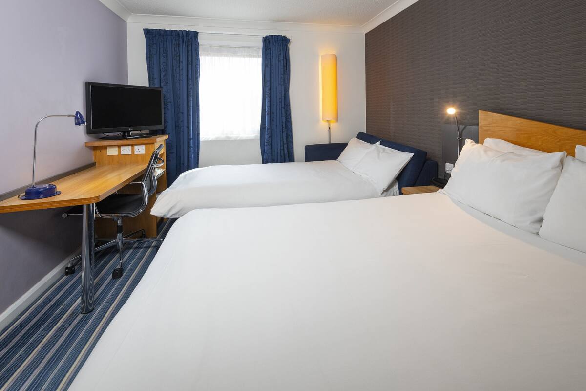 Une chambre d’hôtel avec deux lits, un bureau avec une télévision, une lampe et une fenêtre avec des rideaux.