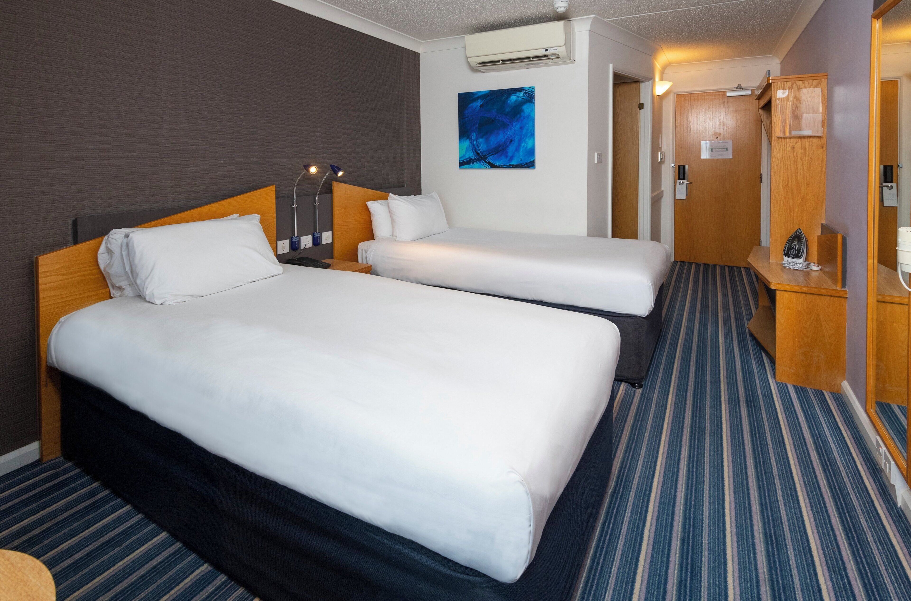 Ett hotellrum med två sängar, en sänggavel i trä, en blå abstrakt målning, en luftkonditioneringsanläggning och en randig matta.