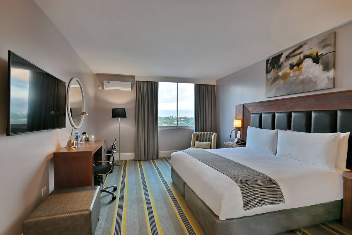 Habitación de hotel con cama, escritorio, silla, televisión, ventana con cortinas y un cuadro en la pared.