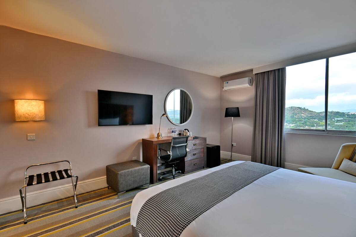 Habitación de hotel con cama, escritorio, silla y un amplio ventanal con vistas.