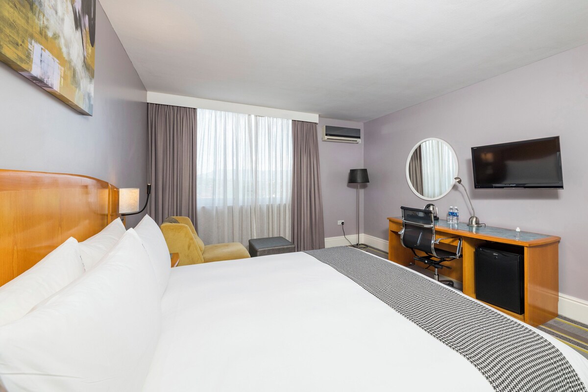 Une chambre d’hôtel comprenant un lit, un bureau avec une chaise, une télévision, un miroir et une fenêtre avec des rideaux.