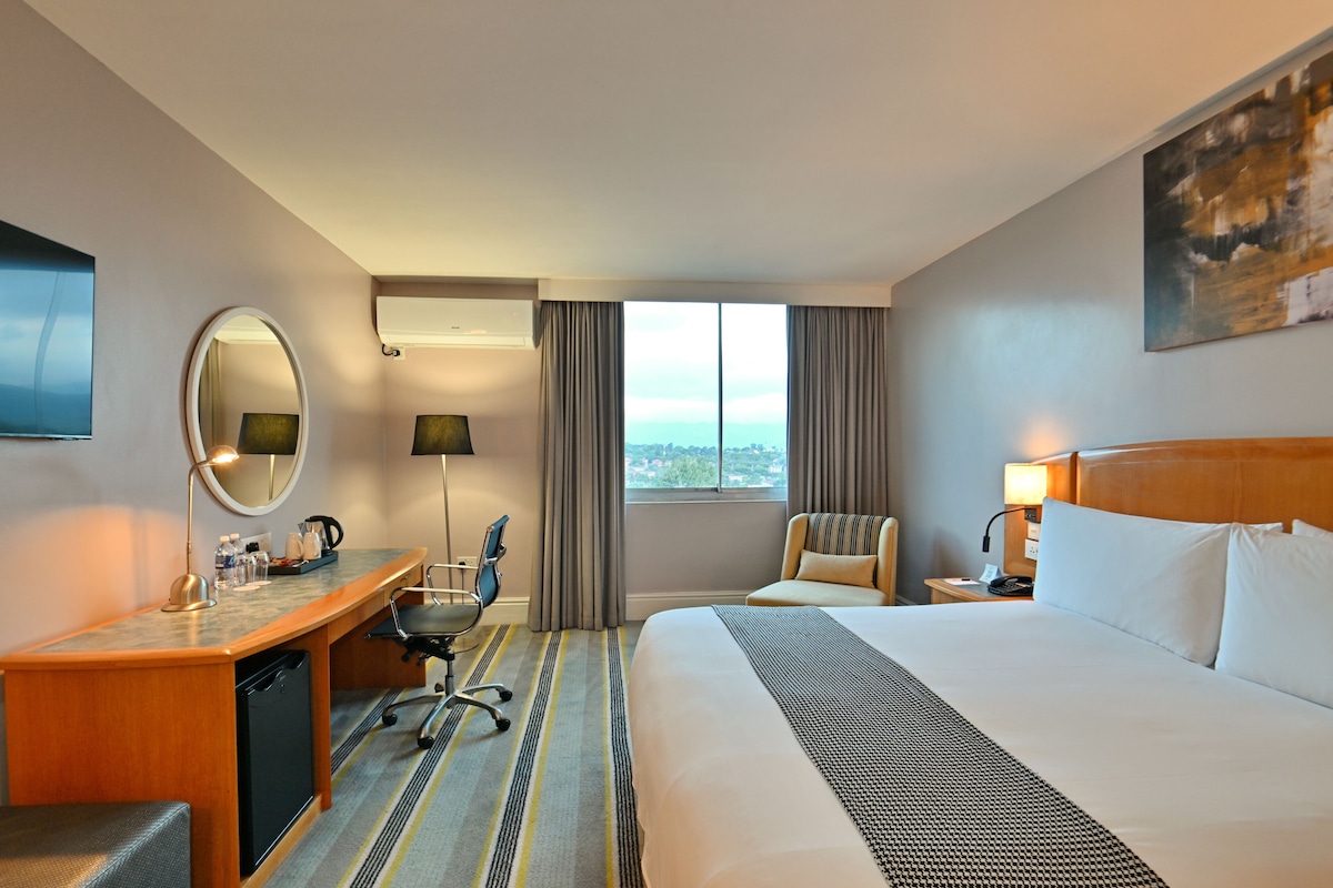 A hotel room with a bed, desk, chair, and a view of the city.