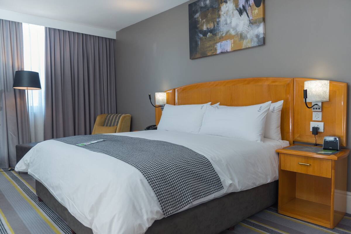 Habitación de hotel con una cama grande, una mesita de noche con lámpara, una silla y un cuadro en la pared.