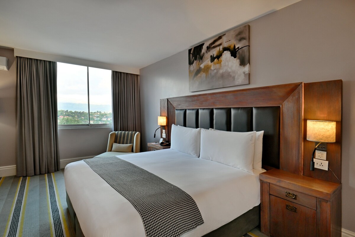 A hotel room with a large bed, a headboard, a nightstand with a lamp, a chair, and a view of the outdoors.