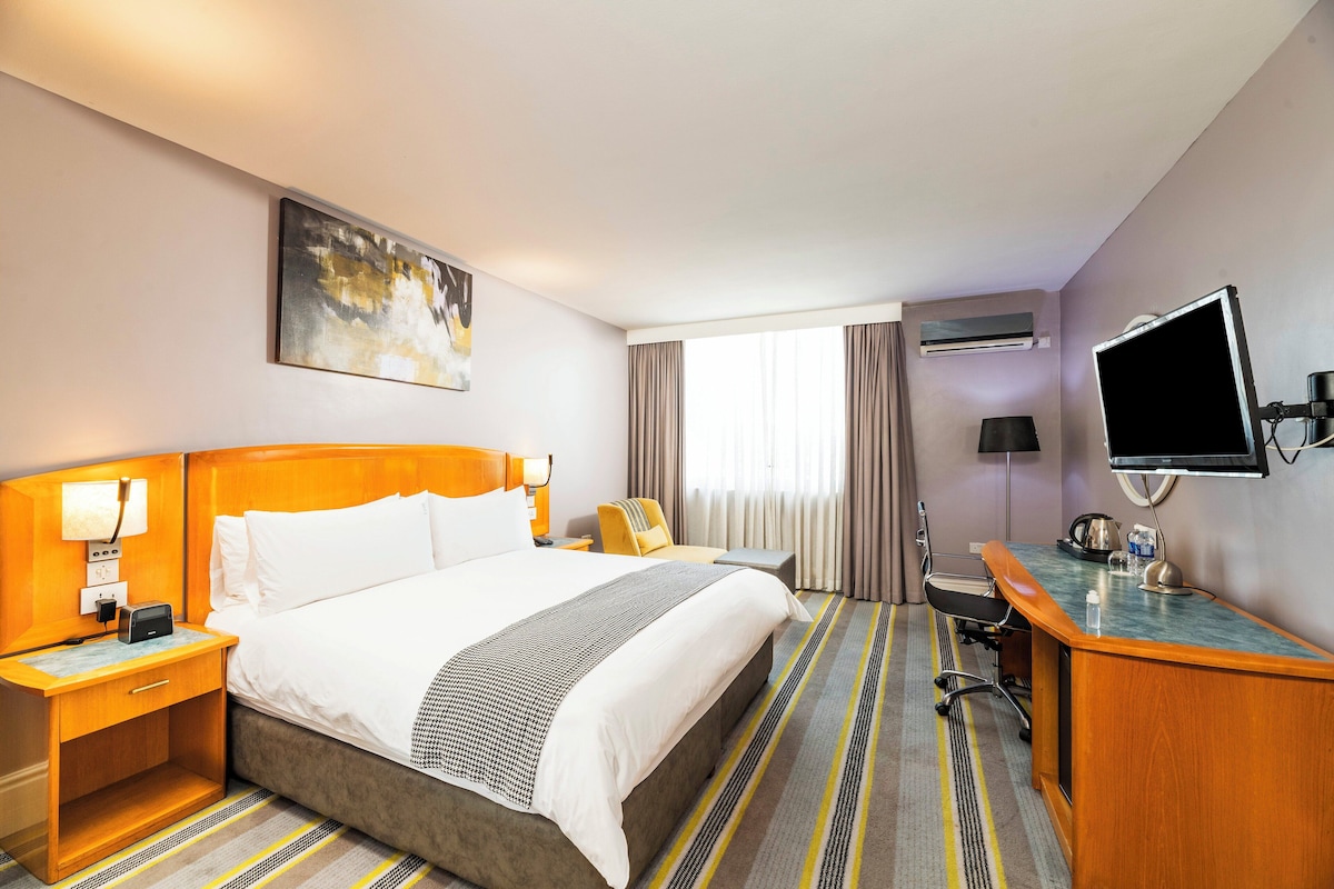 A hotel room with a large bed, a desk, a chair, an air conditioning unit, a television, and a painting on the wall.