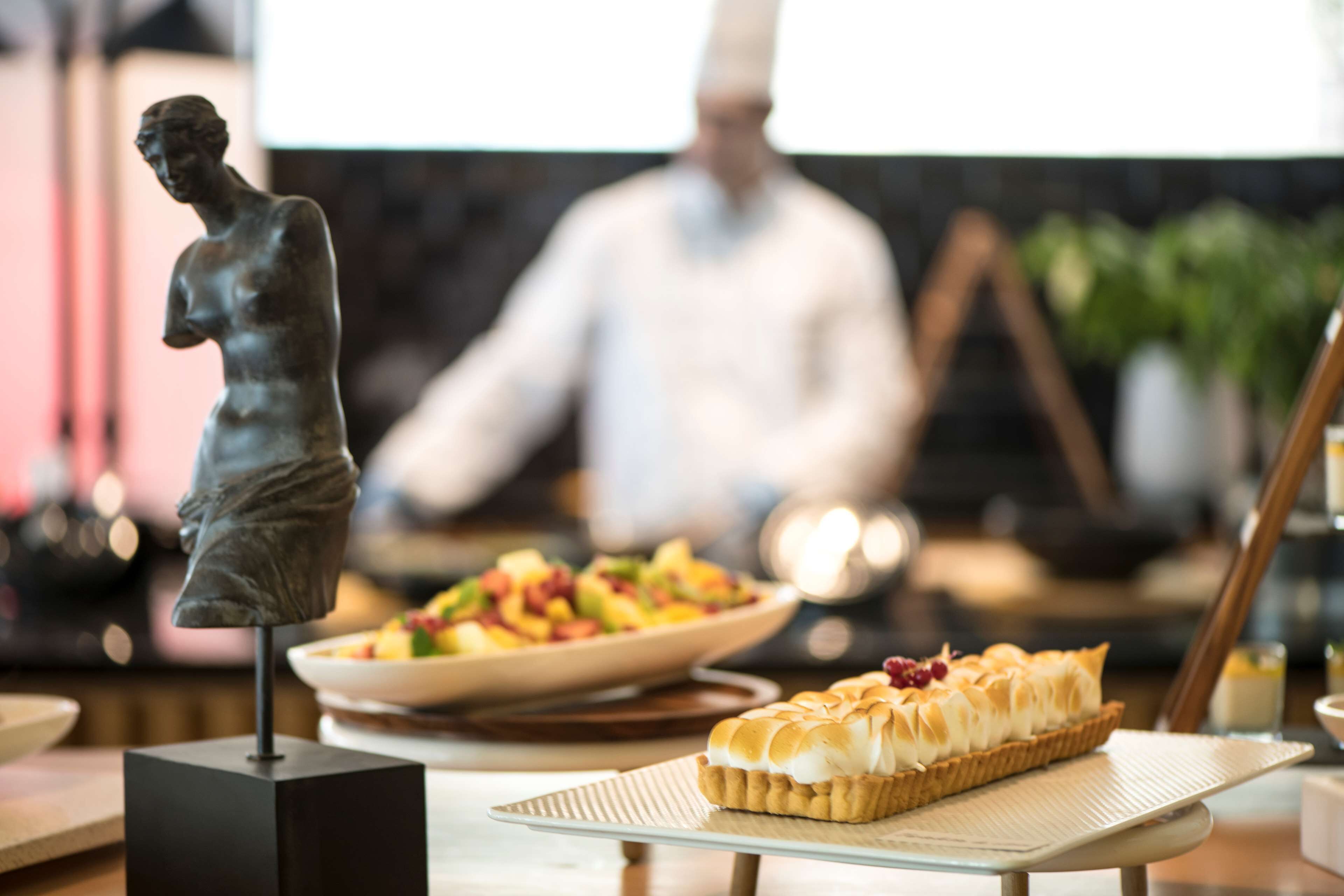 A statue on a pedestal, a dessert on a plate, and a chef in the background.