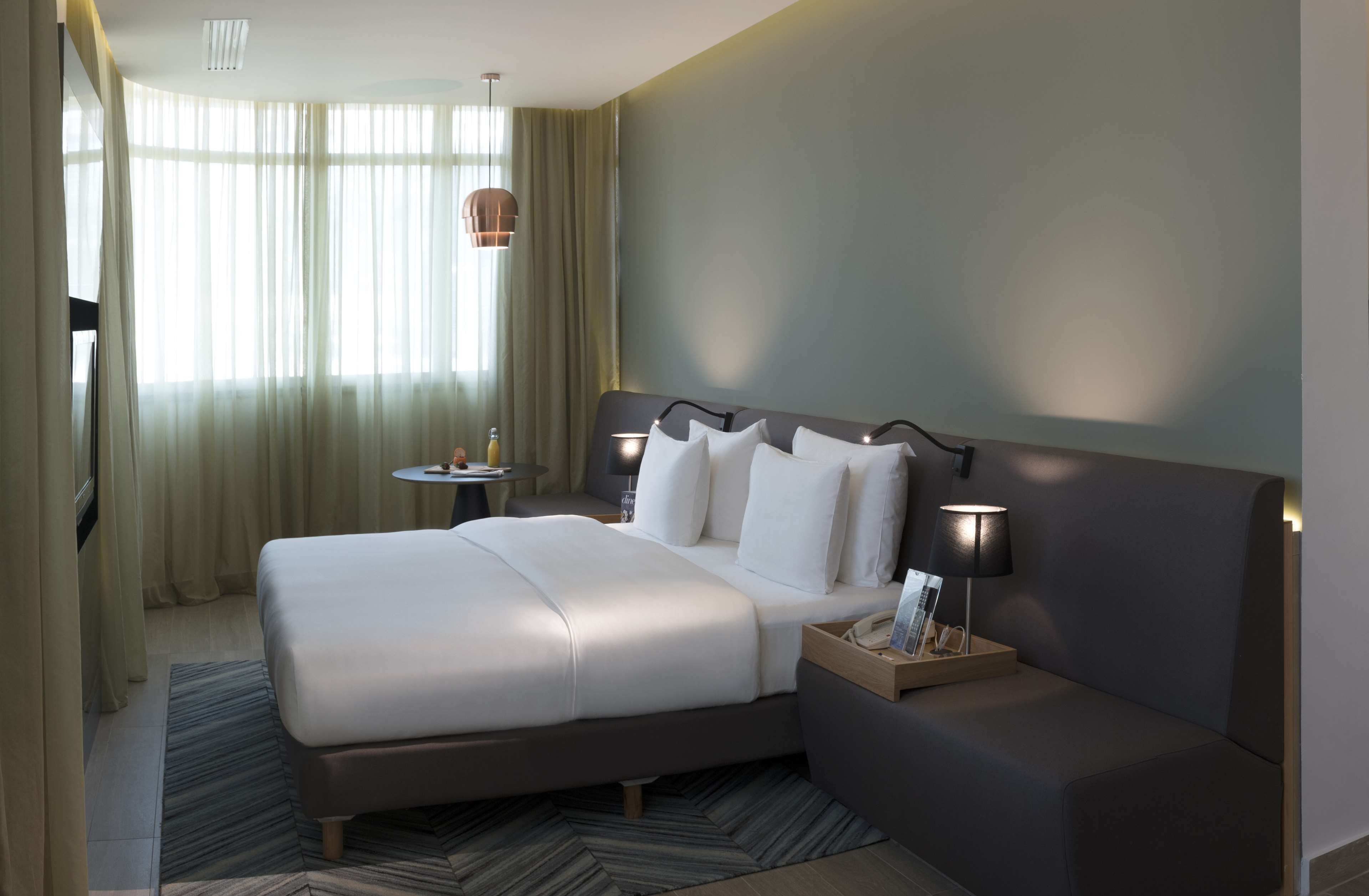 A modern hotel room with a large bed, bedside table, lamp, and a view of the outdoors through sheer curtains.