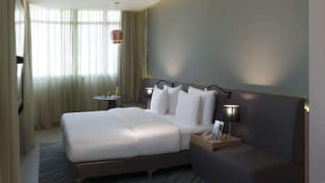 A modern hotel room with a large bed, bedside table, lamp, and a view of the outdoors through sheer curtains.