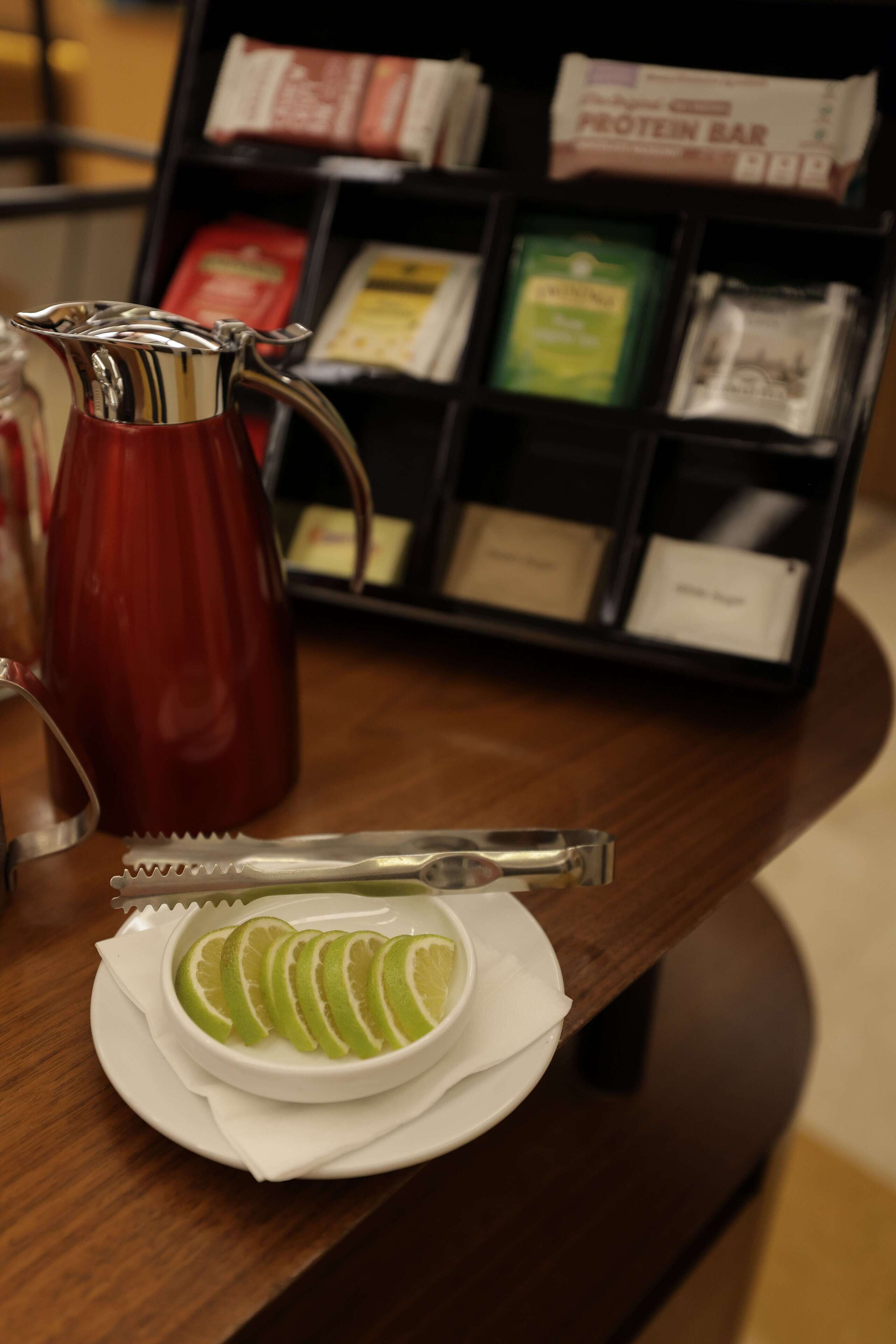A table with a white bowl containing sliced green fruit, a silver pitcher, and a spoon. In the background, there is a shelf with various packaged items.
