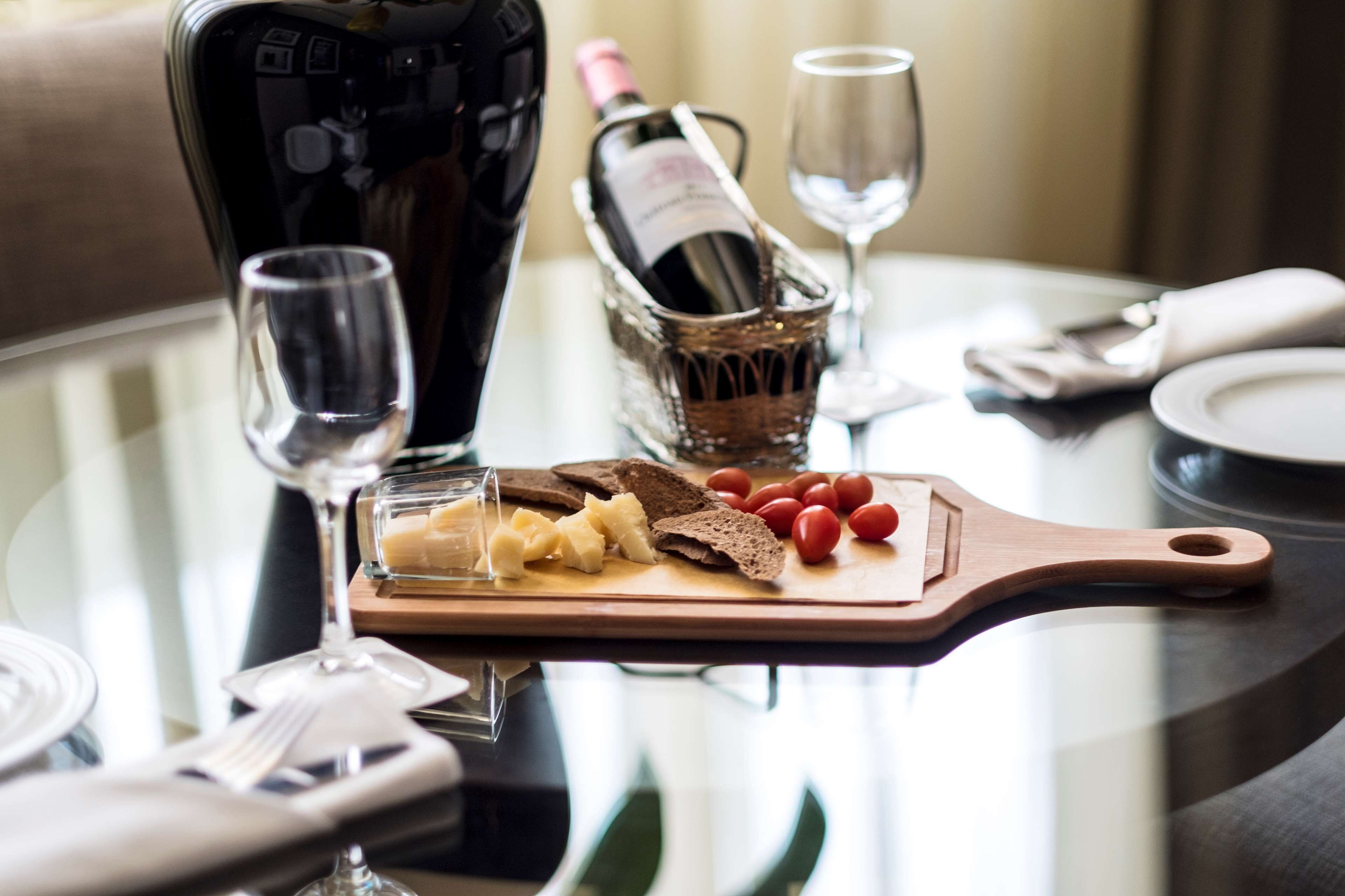Una mesa de comedor con una botella de vino, copas, una tabla de madera con queso y tomates, y una botella de champán.