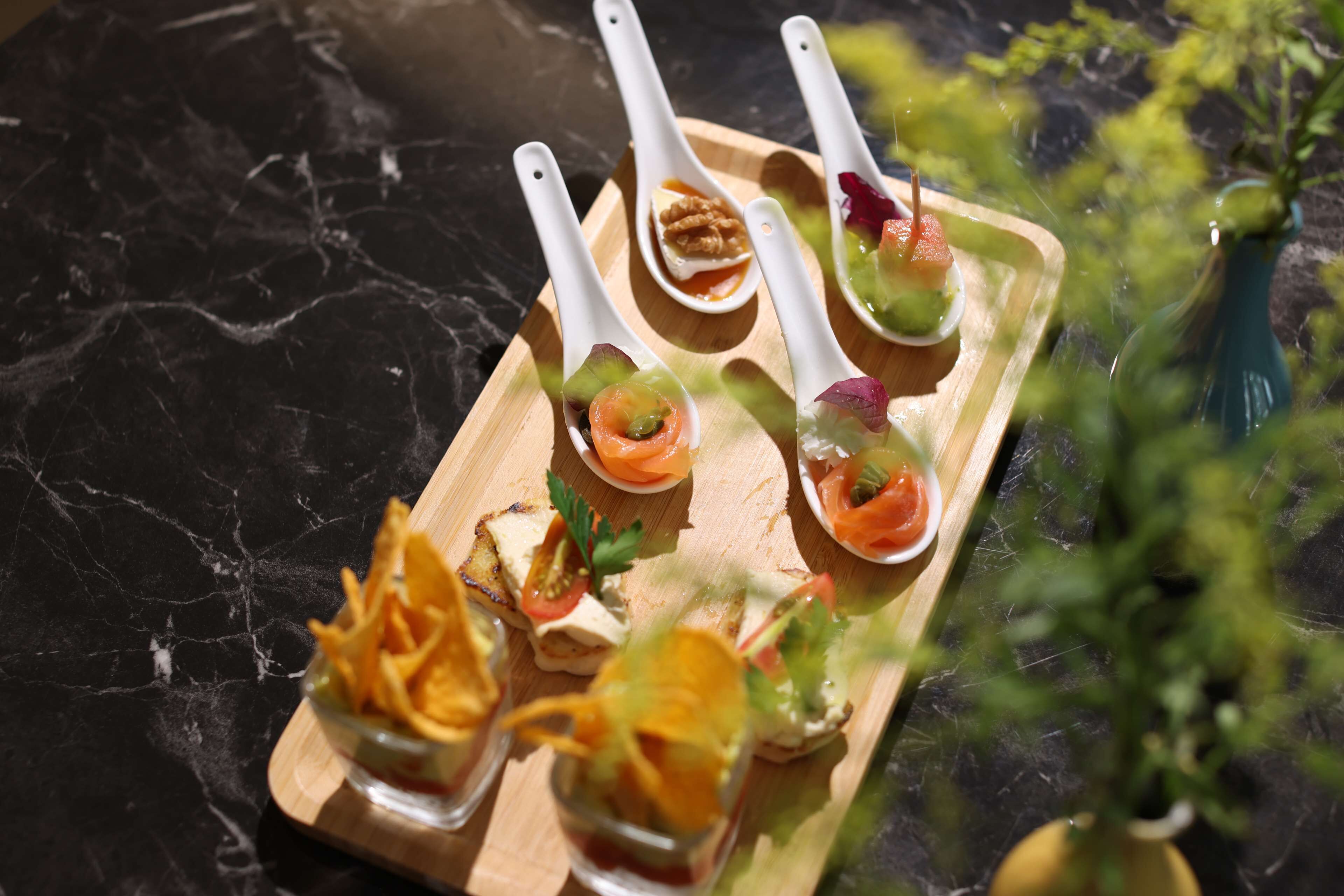 Una bandeja de madera con diversos alimentos, incluyendo un bol de papas fritas, un vaso con ensalada y una cuchara con un trozo de salmón.