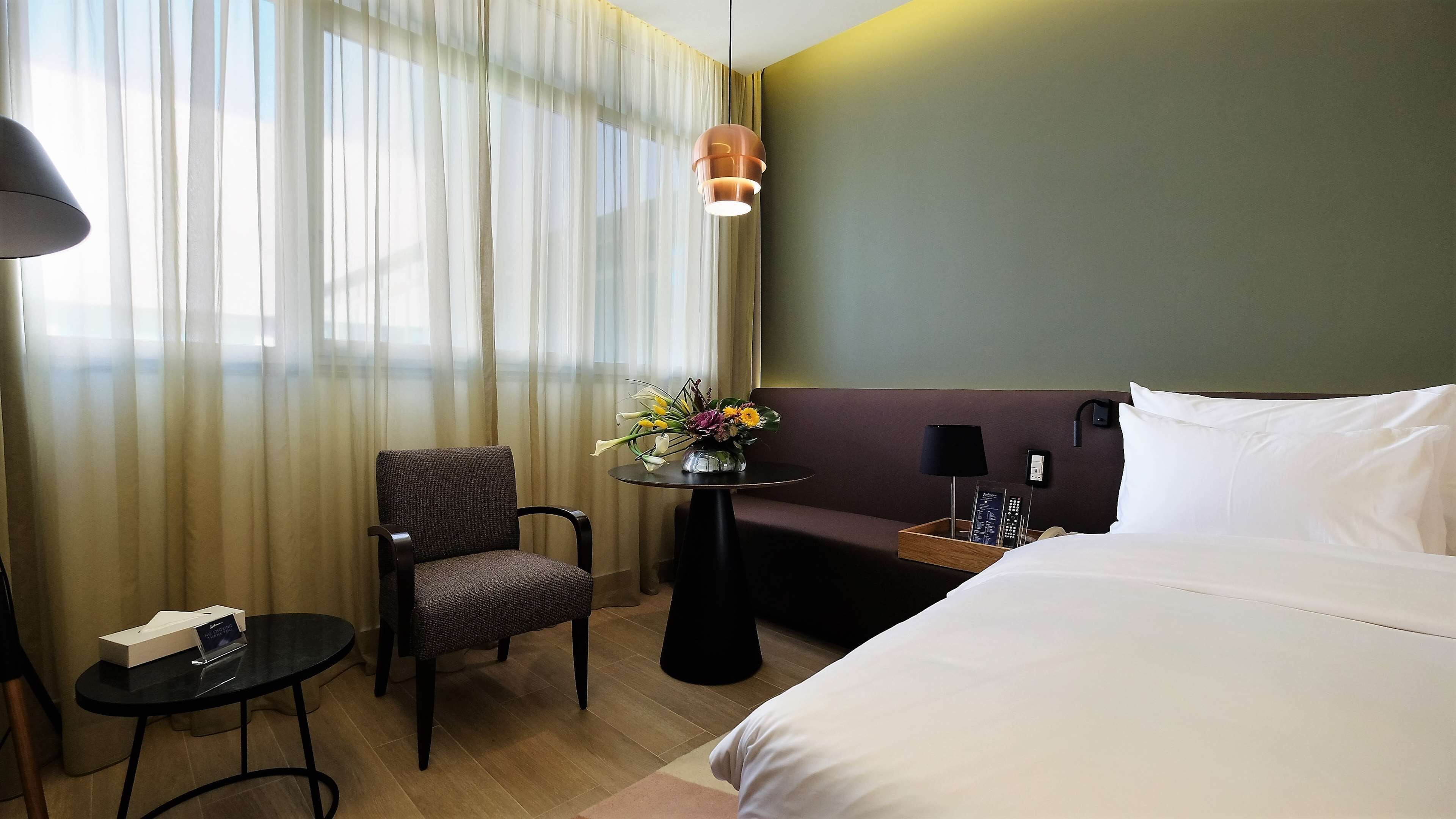 Una habitación de hotel con una cama, una silla, una mesita con un florero, una lámpara y una mesita de noche con un teléfono y controles remotos.