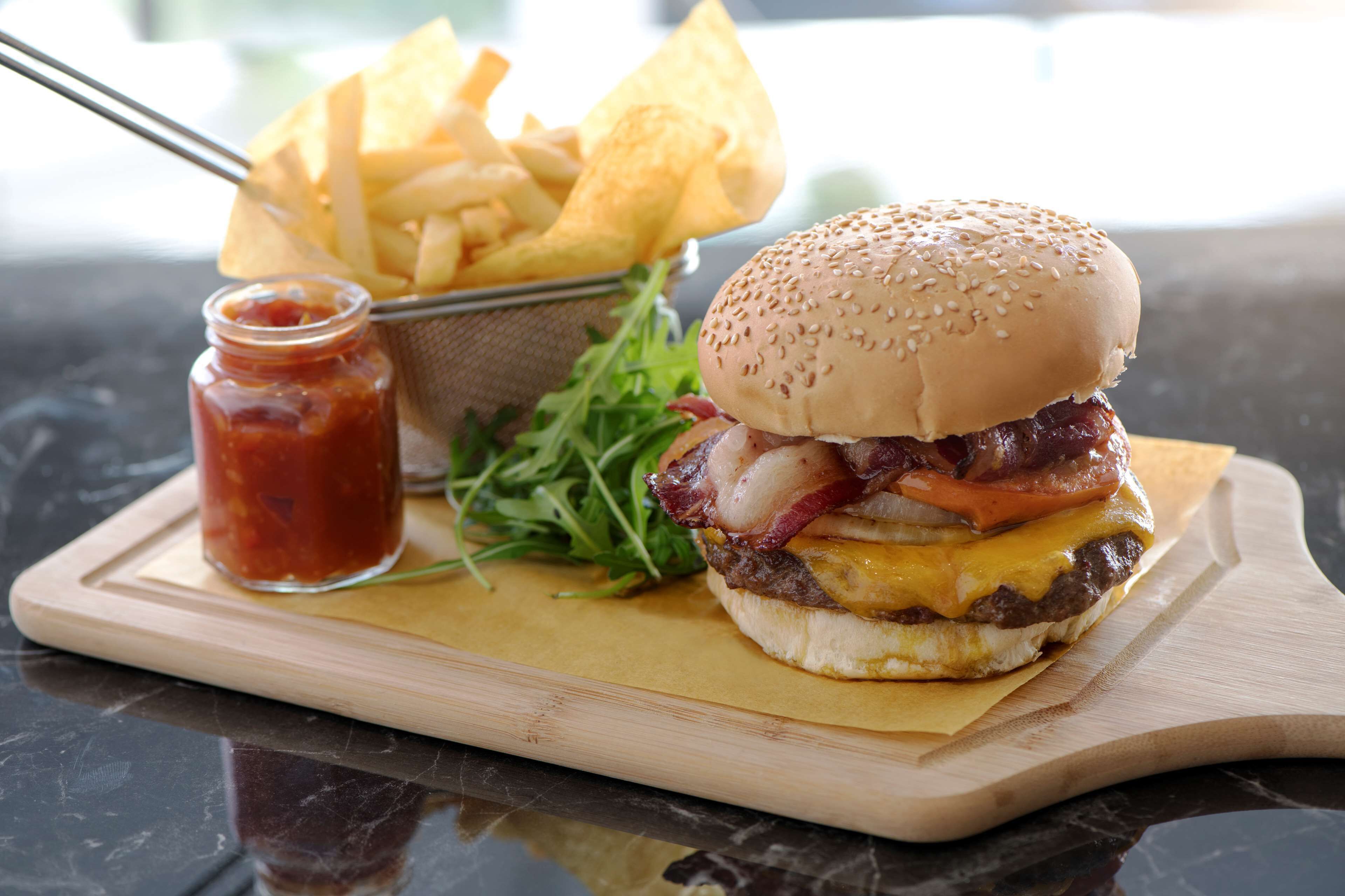 Una hamburguesa con tocino, queso y tomate sobre un pan de sésamo, acompañada de papas fritas y kétchup en una tabla de madera.