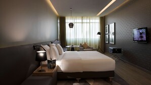 A modern hotel room with a large bed, bedside tables, a desk, a chair, a TV, and a window with curtains.
