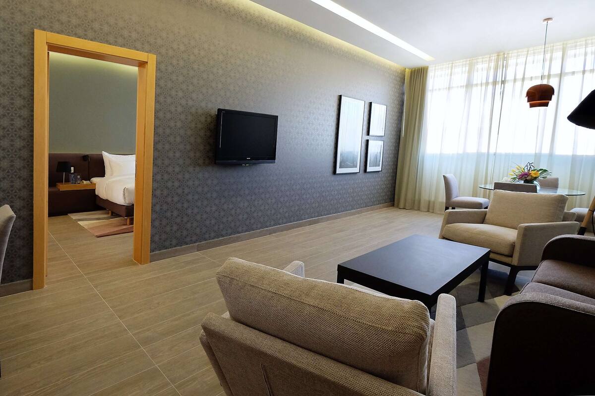 Une chambre d’hôtel moderne avec une porte en bois donnant sur une chambre à coucher, un téléviseur à écran plat fixé au mur et un coin salon avec une table basse et des fauteuils.