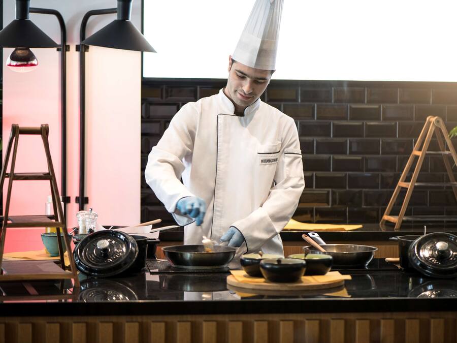 Un cocinero con uniforme y gorro blanco prepara comida en una barra, con sartenes negras, una escalera y un fondo de pared de ladrillo.