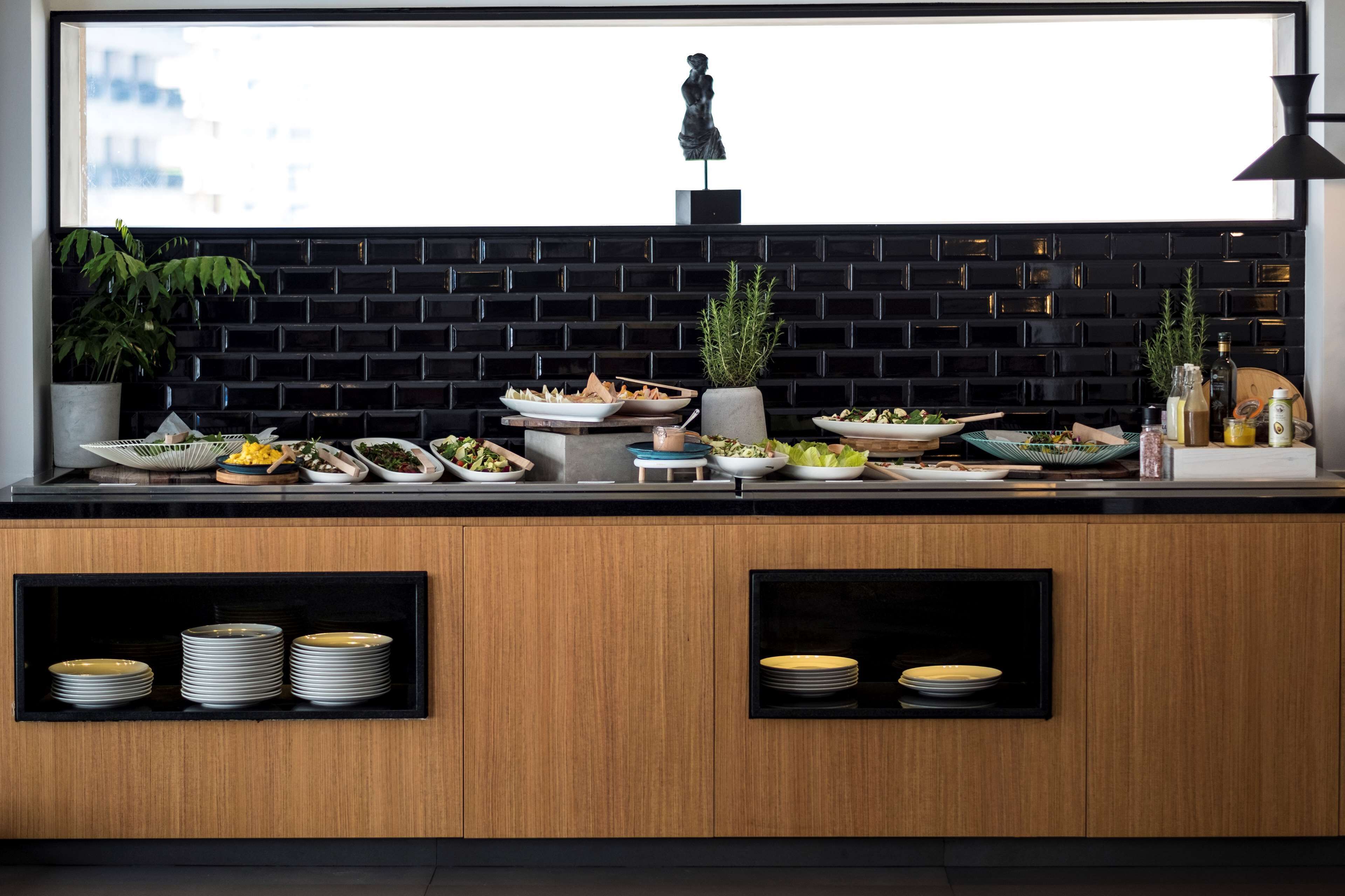A buffet setup with various dishes and drinks, a statue, and a window with a city view.