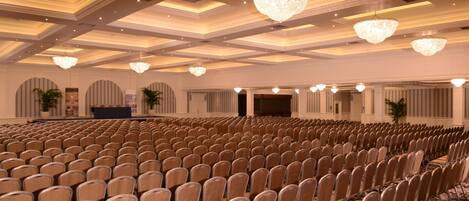 Un salón de conferencias amplio con filas de sillas, candelabros y un escenario.