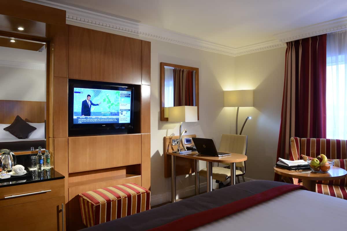Habitación de hotel con cama, escritorio con una computadora portátil, televisión mostrando un noticiero, una silla y una mesita con frutas.
