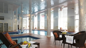 Un área de piscina interior con sillones de descanso y una mesa con un frutero y periódicos.