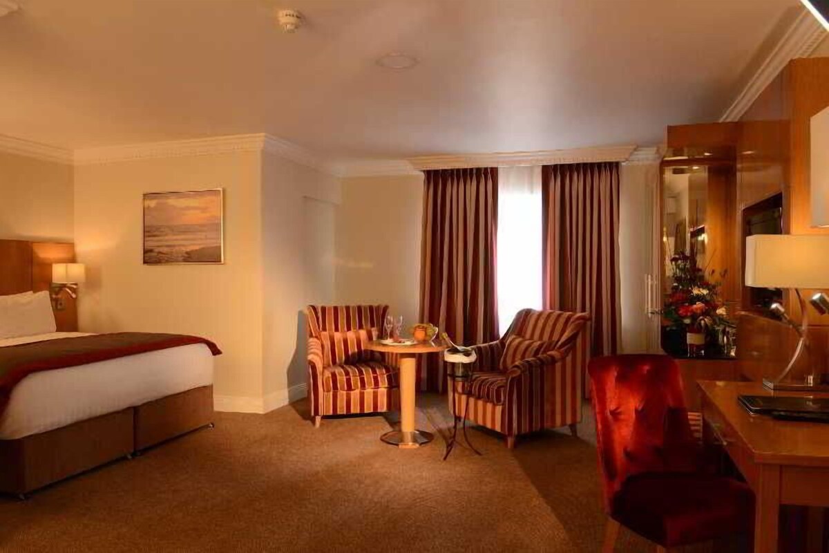 Une chambre d’hôtel avec un lit, deux fauteuils à rayures, une petite table ronde avec une lampe et un bureau avec une chaise.
