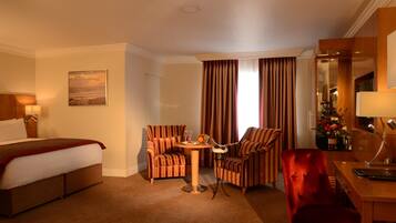 A hotel room with a bed, two striped armchairs, a small round table with a lamp, and a desk with a chair.
