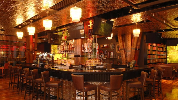 Zona de bar con suelo de madera, taburetes y una variedad de botellas y vasos.