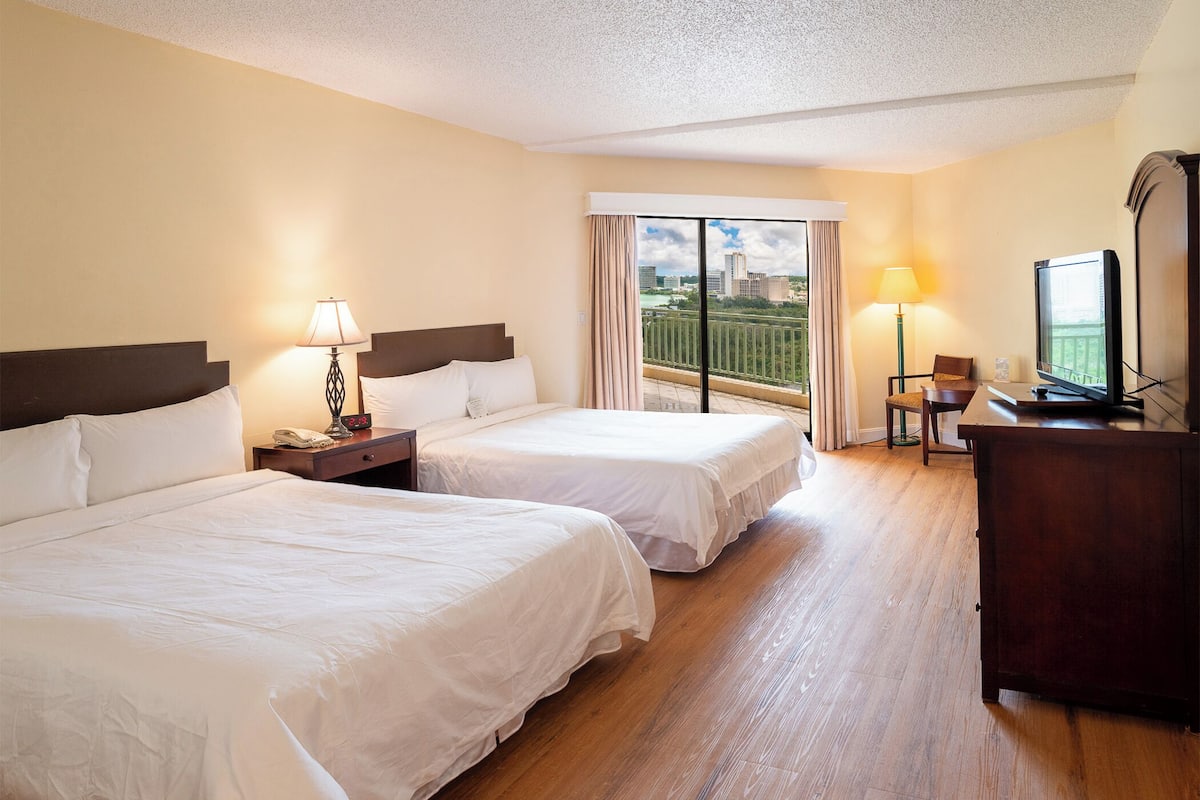 A hotel room with two beds, a desk, a chair, a TV, and a balcony with a city view.