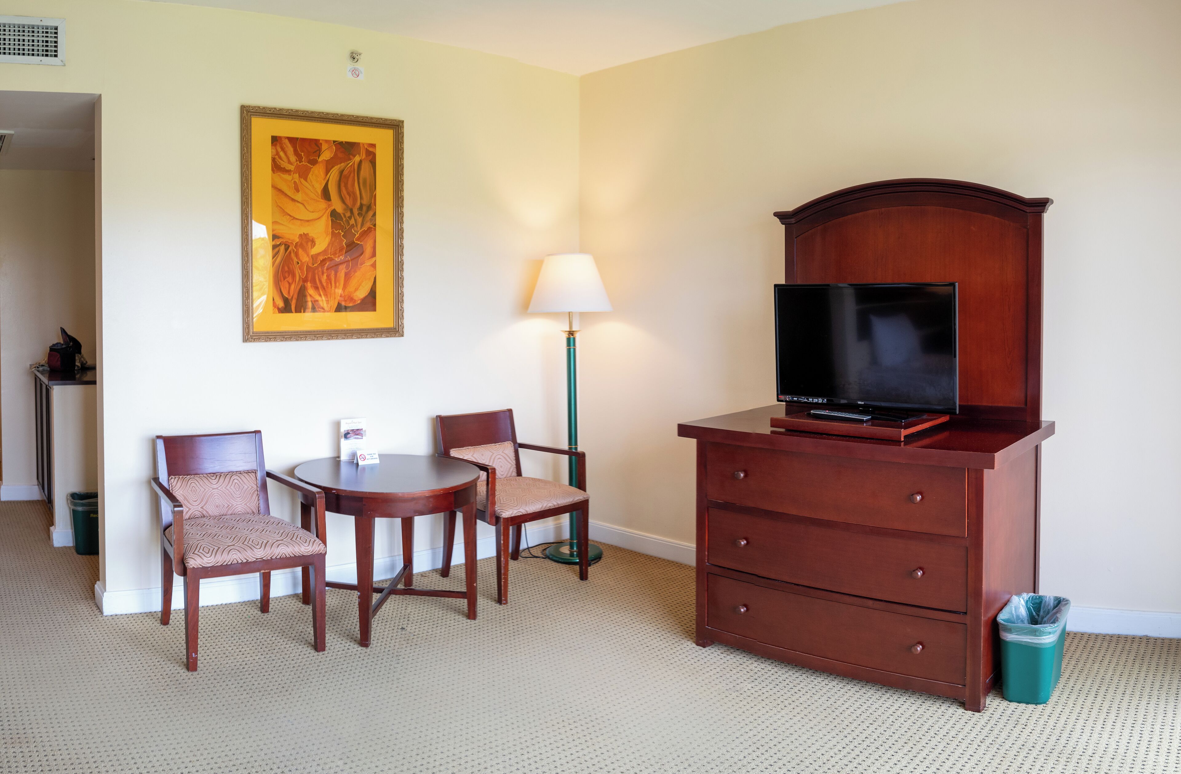 Habitación de hotel con un escritorio de madera, dos sillas, un televisor sobre un mueble de madera, una lámpara y un cuadro en la pared.