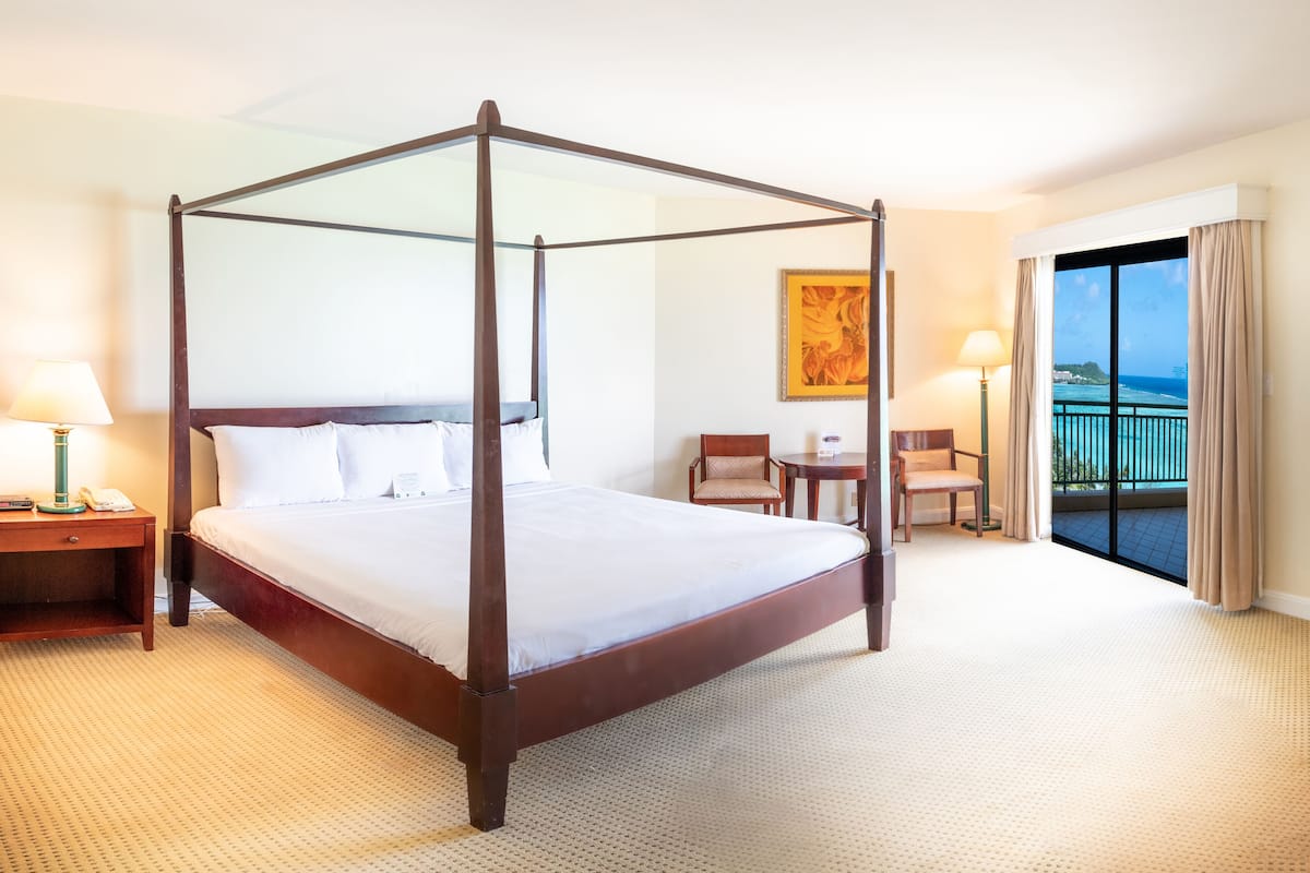 A four-poster bed in a hotel room with a view of the ocean through a balcony.