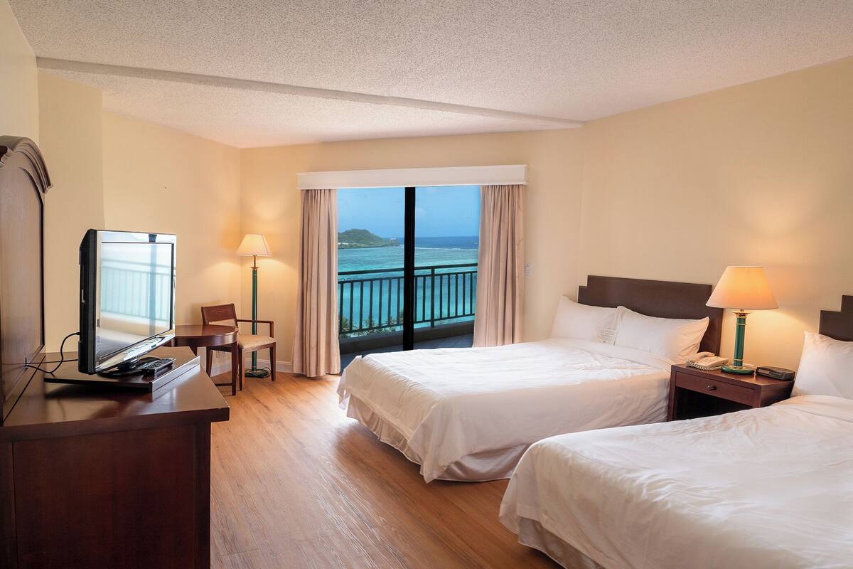 A hotel room with two beds, a desk with a TV, a chair, and a balcony with a view of the sea.
