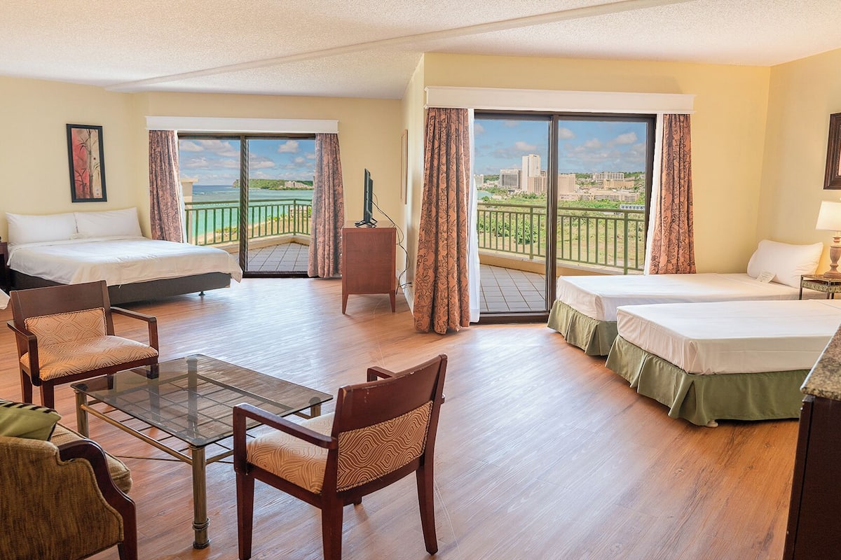 A hotel room with two beds, a sofa, a glass table, and a balcony with a view.