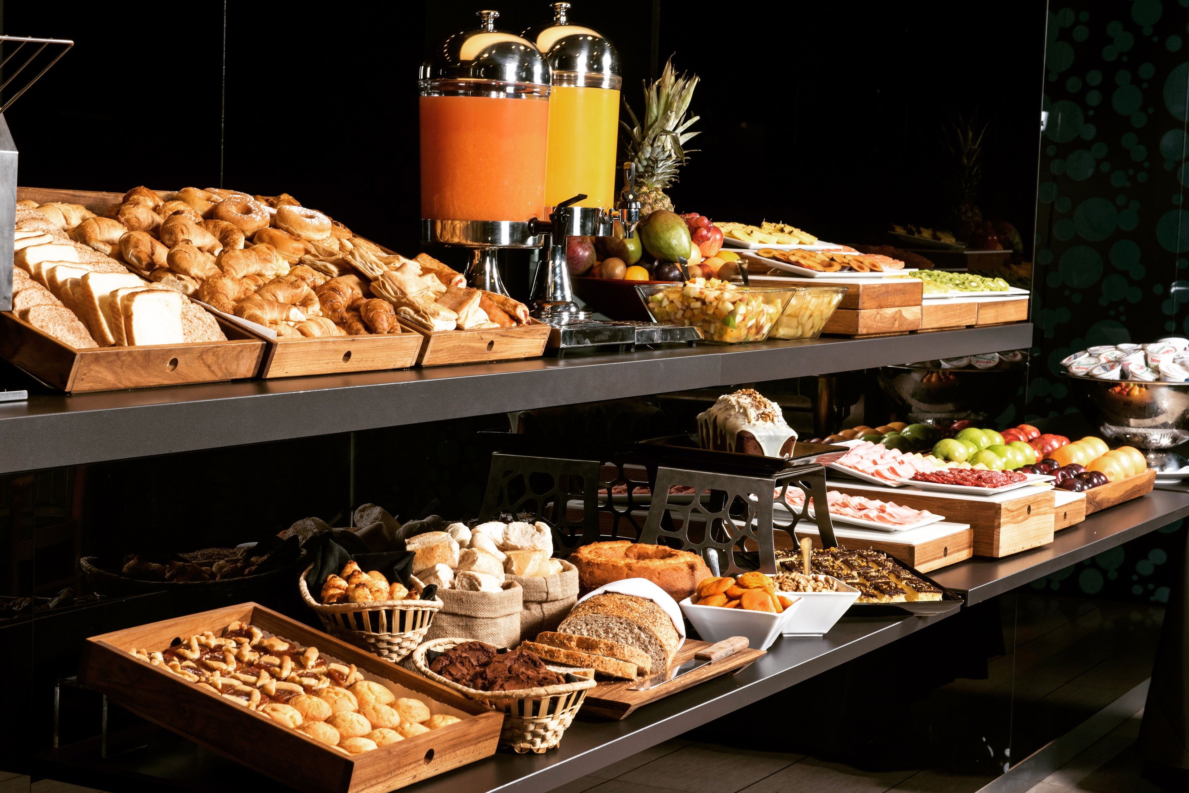 A buffet with various food items including bread, pastries, fruits, and a juice dispenser.